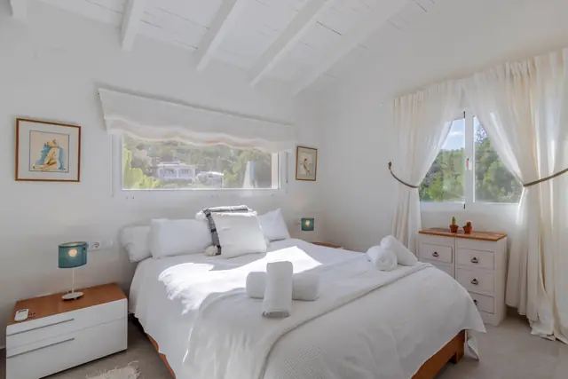 Een slaapkamer met een tweepersoonsbed, een wit balkenplafond en een laag raam met uitzicht op het groen.
